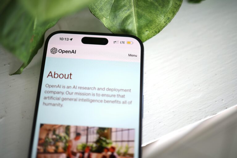 Close-up shot of a smartphone screen showing the OpenAI website with greenery in the background.